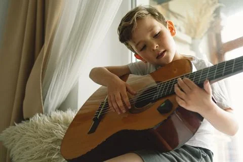 Cute boy learns to play the classical guitar on the windowsill near the window. Stock Photos