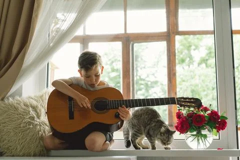 Cute boy learns to play the classical guitar on the windowsill near the window. Stock Photos