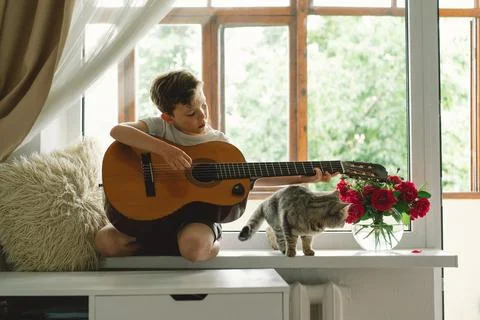 Cute boy learns to play the classical guitar on the windowsill near the window. Stock Photos