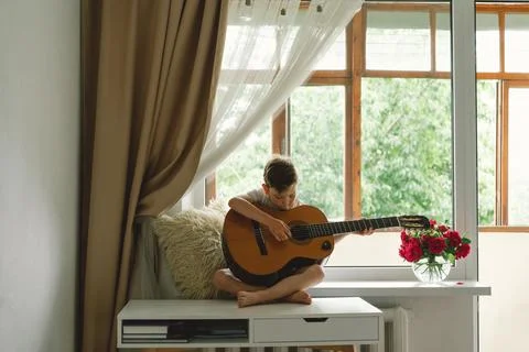 Cute boy learns to play the classical guitar on the windowsill near the window. Foto stock