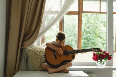 Cute boy learns to play the classical guitar on the windowsill near the window. Stock Photos
