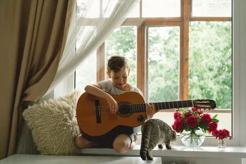 Cute boy learns to play the classical guitar on the windowsill near the window. Stock Photos