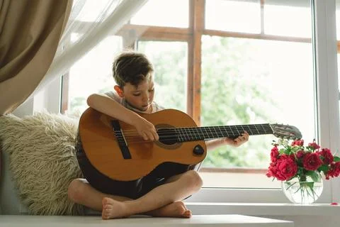 Cute boy learns to play the classical guitar on the windowsill near the window. Stock Photos