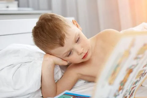 Cute boy lying in a white bed reading a magic book Stock Photos