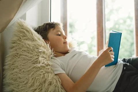 Cute boy reading a book on the windowsill near the window. Stock Photos