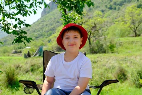 A cute boy in red cap is sitting under the tree and relaxing on mountain back Stock Photos