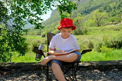 A cute boy in red cap is sitting under the tree and writing in notebook on mo Stock-Fotos