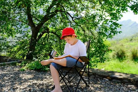 A cute boy in red cap is sitting under the tree and writing in notebook on mo Stock Photos