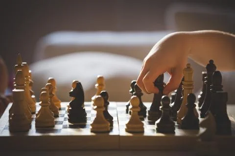 Cute boy sits at a table and plays chess. The kid picked up the chess piece o Stock Photos