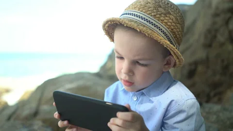 Cute boy sitting on a stone, using smartphone and looking at phone app near sea Stock Footage 106158466