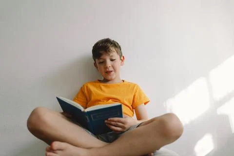 A cute boy wearing an orange T-shirt is sitting on a soft ottoman reading a book Stock Photos