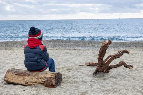Cute boy in winter jacket and knitted hat siting from behind on deserted beach Stock Photos