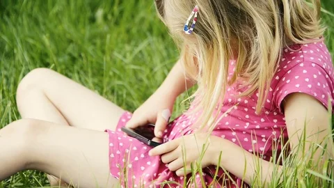 Cute child blond girl sitting in green grass playing with mobile  smart phone Stockbeeldmateriaal 80123341