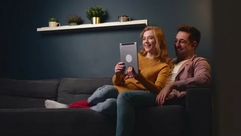 Cute couple are using a tablet sitting on a couch and smile while relaxing Stock Footage 148403563