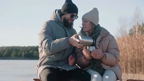 Cute couple drinking hot tea from thermos sitting on a dock in winter near lake Stock Footage 170162081