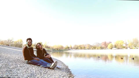 Cute couple sitting by lake and listening to music with headphones Stock Footage 59555323