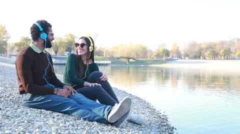 Cute couple sitting by lake and listening to music with headphones Stock Footage 59555330