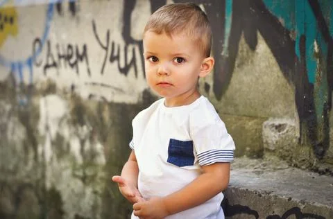 A cute fair-haired boy in a white T-shirt stands modestly and looks fearfully in Stock Photos
