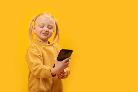 Cute fair-haired kid with two ponytails in yellow jacket looks at phone and s Stockfoto's