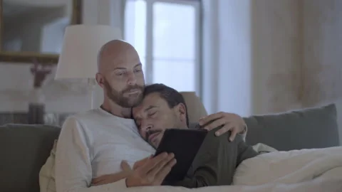 Cute gay lying in bed with partner and reading news on tablet Stock Footage 229630640