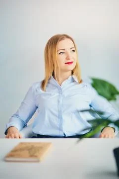Cute girl in the office sits at the table. Young woman in blue shirt smiles and Stock Photos