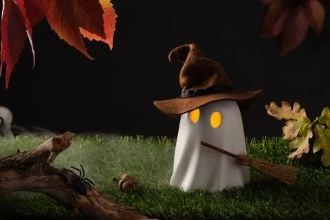 Cute Halloween ghost with witch hat and broom, glowing eyes on green grass wi Stock Photos
