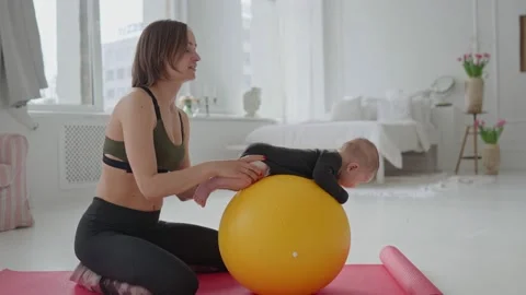 Cute infant boy doing exercises lying on fitness ball while mother holding her Stock Footage 151346423