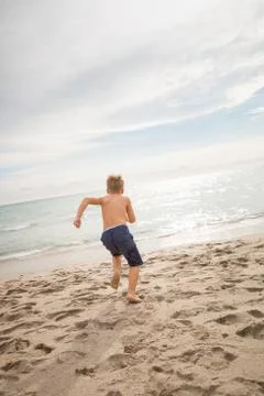 Cute kid boy running fast into the ocean on the sand beach. Clouds background Stock Photos