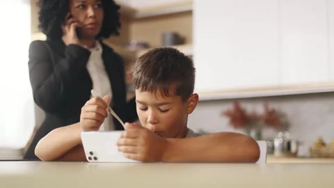 Cute kid with breakfast hold smartphone while busy mother has working call Stock Footage 248689702