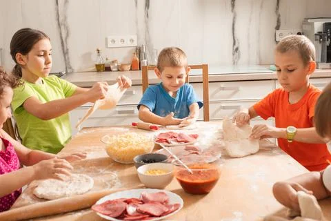 The cute kids sit at a table in the kitchen and make pizza Stock Photos