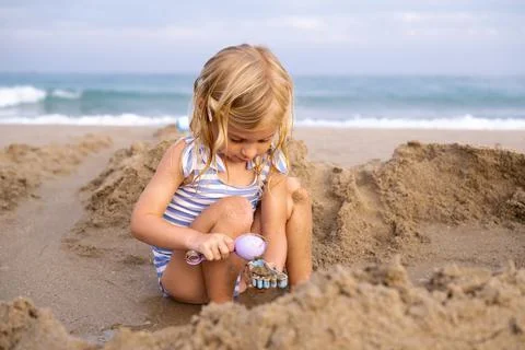 Cute laughing blonde three years old girl playing with sand at beach near sea in Stock Photos