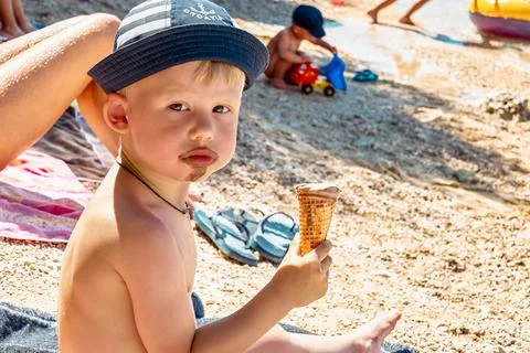 Cute little boy in hat eats ice cream sitting on beach Stock Photos