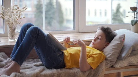 Cute little boy lying on windowsill and reading book Vídeos de archivo 104243883