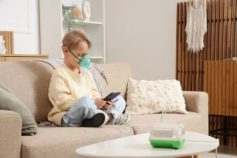 Cute little boy with nebulizer using mobile phone on sofa at home Stock Photos