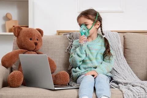 Cute little girl with laptop using nebulizer on sofa at home Stock Photos