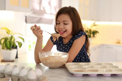 Cute little girl licking raw dough from spoon in kitchen. Cooking food Stock Photos