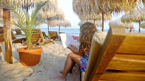 Cute little girl reading book among straw parasols and seabeds on the beach Stock Footage