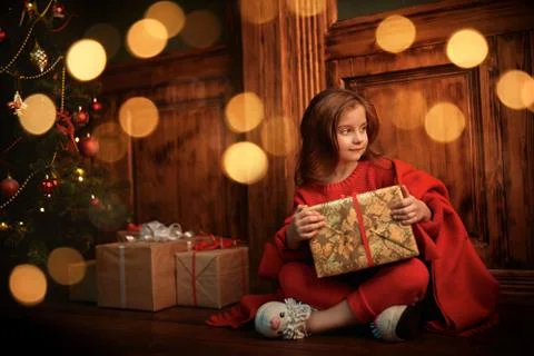 Cute little girl sits with a gift box by a beautiful Christmas tree. Christma Stock Photos