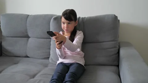 Cute little kid sitting on couch watches TV with interest, holding tv remote in Stock Footage 218237180