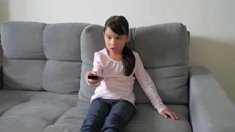 Cute little kid sitting on couch watches TV with interest, holding tv remote in Stock Footage 219024938