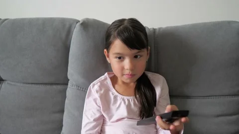 Cute little kid sitting on couch watches TV with interest, holding tv remote in Stock Footage 219944847