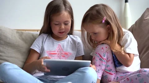 Cute little sisters sitting close to each other, watching something on tablet Видео 139208945