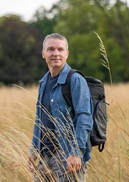 Cute middle aged man with backpack standing in tall dry grass and enjoying pe Stock Photos