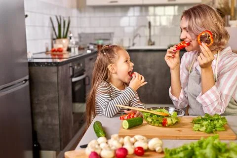 Cute mom and child girl tasting fresh fruits while preparing salad for dinner Stock Photos