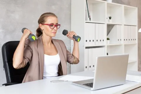 Cute office worker doing exercises with dumbbells and looking at laptop Stock Photos