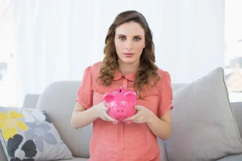 Cute pregnant woman holding a piggy bank while sitting on couch Stock Photos