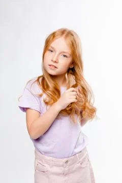 Cute red-haired long-haired girl in daily clothes (skirt, T-shirt) braids her Stock Photos
