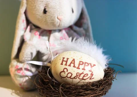 A cute sad toy bunny on blue background and a nest with an Easter egg inside. Stock Photos