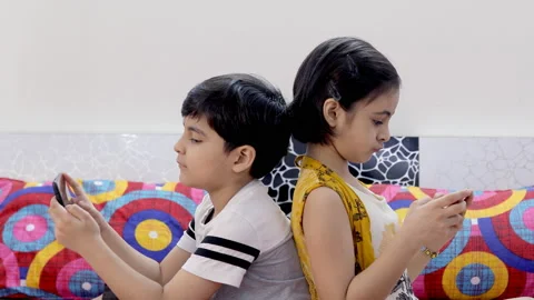 Cute siblings playing games on their mobiles while sitting together in the be... Vidéo 139099885