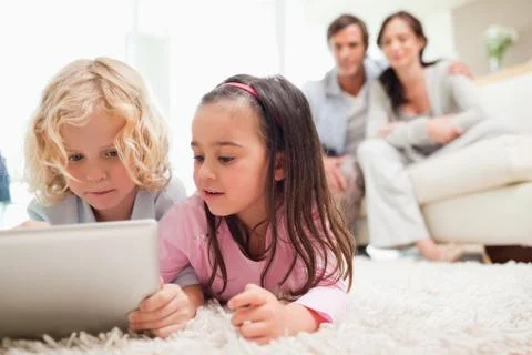 Cute siblings using a tablet computer while their parents are in the background Stock Photos